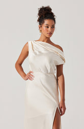 A woman is wearing an elegant off-the-shoulder satin dress in a light cream color. The dress features a draped neckline and a high slit on the side, revealing part of her leg. The fabric has a smooth, shiny finish, giving it a luxurious appearance.